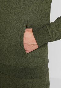 Olivově zelená mikina s texturovaným povrchem, vybavená boční kapsou a žebrovanými manžetami pro přiléhavý vzhled.