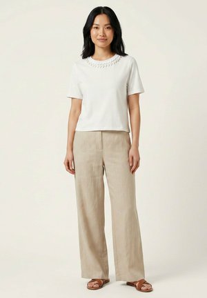 Femme aux longs cheveux foncés portant un haut blanc à manches courtes et un pantalon beige à jambes larges avec des sandales marron sur un fond uni.