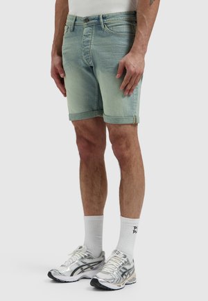 Man draagt lichtblauwe denim shorts met opgerolde zoom, witte crew-sokken en zilveren hardloopschoenen, staand tegen een effen achtergrond.