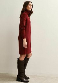 Robe en tricot avec col montant de couleur rouge profond, à manches longues et poignets côtelés, accompagnée de bottes en cuir marron, à talons carrés, hauteur genoux.
