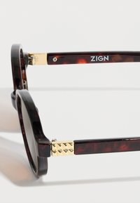 Καφέ γυαλιά ηλίου με στρογγυλούς φακούς, χρυσαφένιες λεπτομέρειες στα πλαϊνά και διακοσμητικά καρφιά στη άρθρωση. Λογότυπο "ZIGN" στο μπράτσο.