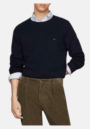 Mężczyzna ubrany w granatowy sweter na białą koszulę w paski zapinaną na guziki oraz brązowe spodnie ze sztruksu, z ręką spoczywającą przy boku i biodrze.