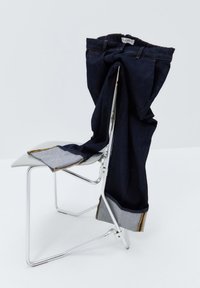 Jeans en denim noir drapés sur une chaise en chrome, avec un revers gris plié et des coutures jaunes, et une étiquette de taille visible sur le dessus.
