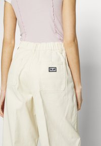 Pantalon de couleur crème avec une taille élastique, doté de deux poches arrière et d'un petit patch logo noir. Le tissu semble doux et texturé.