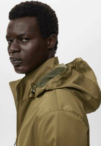 Chaqueta verde oliva con un alto cuello y capucha ajustable, presenta un cierre de cremallera y tejido texturizado. Incluye una costura de hombro prominente.