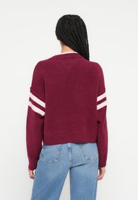 Tumepunane kampsun roosade triipudega varrukatel, soonikkoes äärte ja mansettidega, valmistatud pehmest tekstuuriga kangast. Tagantvaates on näha v-kaelusega disain.