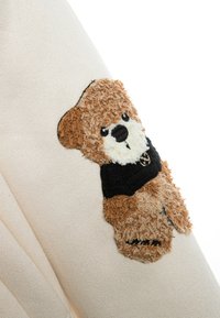 Tela color crema con un diseño texturizado de un oso de peluche bordado en marrón y negro, con un notable detalle parecido al pelo.
