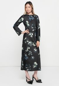 Long black dress with a floral print in shades of green and white, featuring long sleeves and a straight silhouette. Paired with black heeled shoes.