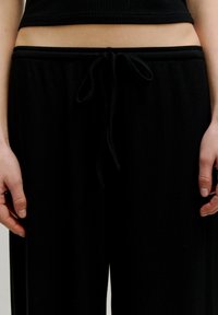 Zwarte geribbelde stoffen broek met een elastische tailleband en trekkoord. De stof heeft een gladde textuur en biedt een losse, relaxte pasvorm.