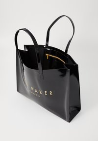 Bolsa tote de verniz preto com letras em dourado e duas alças longas. O interior inclui um bolso com fecho éclair dourado.