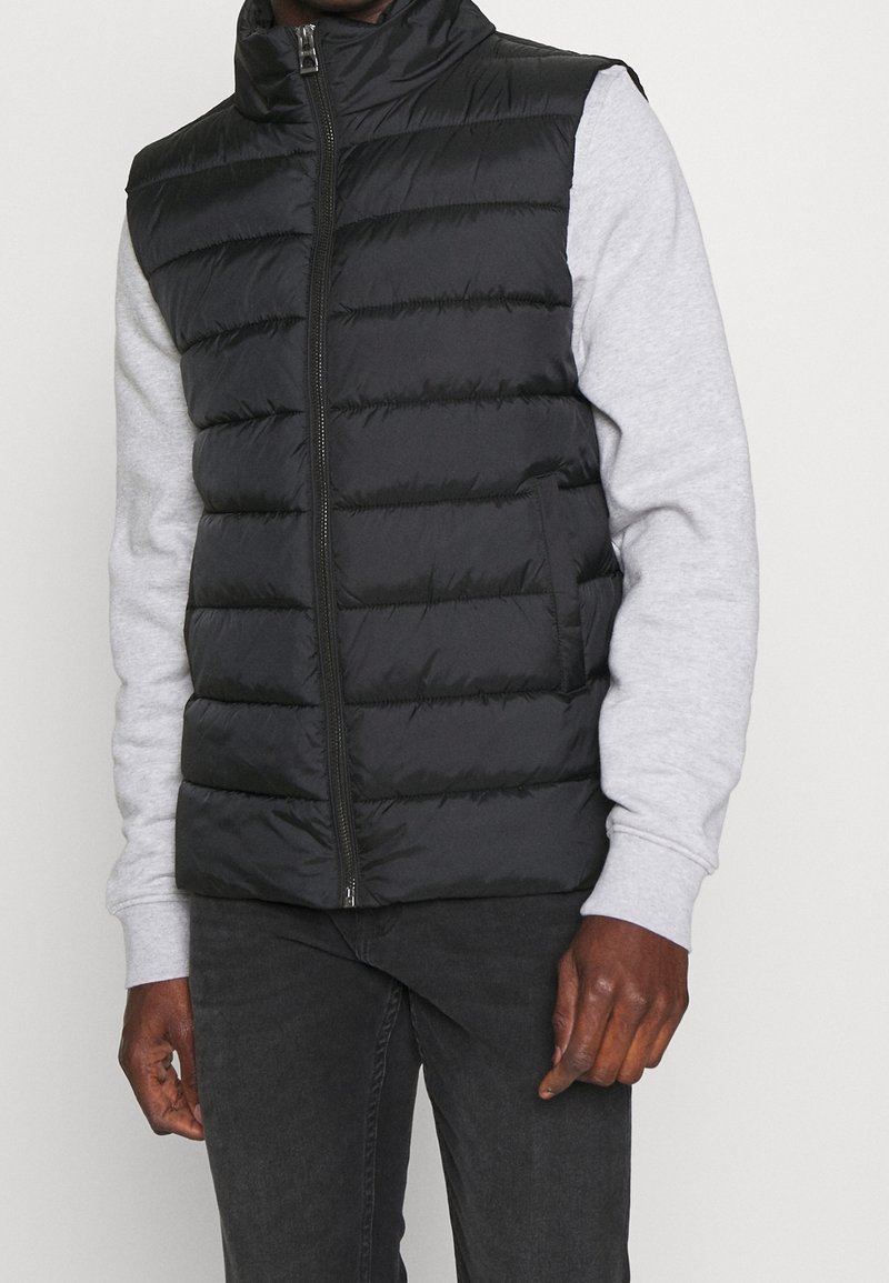 Homme portant un gilet sans manches noir matelassé sur une chemise à manches longues gris clair avec un pantalon foncé, debout devant un fond clair uni.