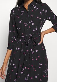 Svart skjortklänning med lila och rosa blommönster, knäppning framtill, krage och knytmidja. Har trekvartsärmar och insvängd midja.