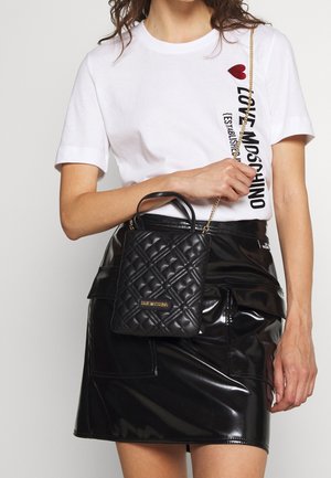 Černá prošívaná kabelka se zlatým logem, zlatým řetízkovým páskem a horními uchy. Modelka má na sobě bílé tričko s potiskem a černou lesklou minisukni.
