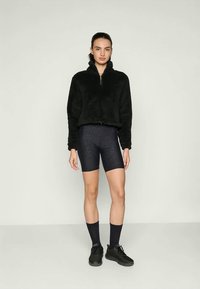 Giacca in pile nera con zip e colletto alto, abbinata a shorts da ciclismo scuri con motivo. Modello con calzini neri e sneakers.