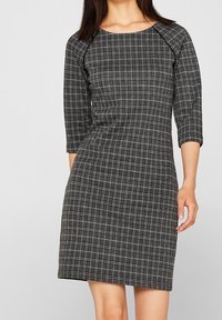 Musta ruudullinen mekko, jossa on tiukka muotoilu, kolme neljännestä hihaa ja pyöreä pääntie. Valmistettu teksturoidusta kankaasta, jossa on valkoisia yksityiskohtia.
