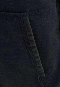 Donkergekleurde stof van dichtbij, met een gestructureerd oppervlak en zichtbare stiksels langs de rand van de zakken. De stof heeft een lichte rek.