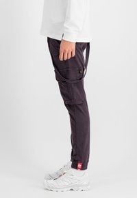 Cargo-Hosen aus dunkelviolettem Stoff, mit mehreren Taschen, einem lässigen Schnitt und einem Kordelzug am Saum, kombiniert mit weißen Sportschuhen.