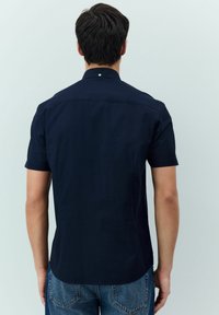 Kniekurzes Hemd aus glattem Stoff in Marineblau, mit umlegbarem Kragen und einem dezenten Nahtdetail im oberen Rückenbereich.