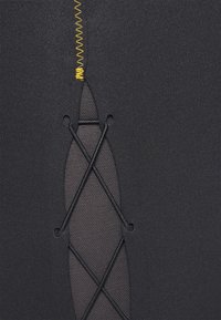 Svart atletisk tyg med texturerad meshpanel och korslagda elastiska detaljer, framhävd av gul zigzag-stickning längs sömmen.