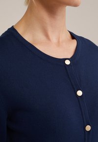 Cardigan blu navy con scollo rotondo, caratterizzato da bottoni dorati sulla parte anteriore. Realizzato in tessuto morbido e texturizzato.