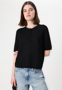 Maglia nera a maniche corte in maglia con scollatura ondulata e chiusura a bottoni; presenta un motivo testurizzato e una vestibilità rilassata. Indossata con jeans chiari.