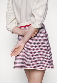 Personne portant une jupe à carreaux rouge, blanc et bleu avec un chemisier blanc à fines rayures, mains jointes dans le dos.
