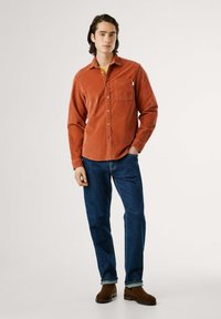 Roestrode corduroy overhemd met knopen en een borstzak, gecombineerd met donkerblauwe spijkerbroek en bruine enkelboots.