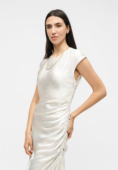 Γυναίκα με μακριά σκούρα μαλλιά, που φοράει ένα αμάνικο λευκό σατέν φόρεμα με ραφές και χρυσό κολιέ αλυσίδα, ποζάροντας με το χέρι στη μέση.