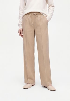 Vrouw draagt beige broek met wijde pijpen en een taille koord, gecombineerd met een crèmekleurige top met lange mouwen en beige platte schoenen op witte achtergrond.
