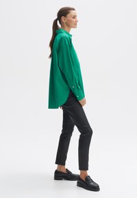 Groene oversized button-up shirt met een puntige kraag, lange mouwen en afgeronde zoom, gecombineerd met zwarte slim-fit broeken en zwarte instappers.