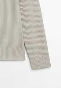 Détail de la manche et de l'ourlet d'un pull en maille côtelée gris clair sur fond blanc.