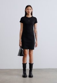 Μαύρο εφαρμοστό φόρεμα με κοντά μανίκια, στρογγυλή λαιμόκοψη και διακριτική λεπτομέρεια με λογότυπο. Συνδυασμένο με μαύρες μπότες μέχρι το γόνατο και μια μικρή τσάντα.