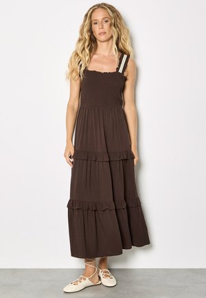 Frau mit langen, lockigen blonden Haaren, die ein ärmelloses, braunes, gestuftes Maxikleid und cremefarbene, flache Schnürschuhe trägt, steht vor einer weißen Wand.