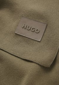 Olivengrønt strikket stof med en struktureret overflade og et brunt lædermærke med det prægede ord "HUGO."
