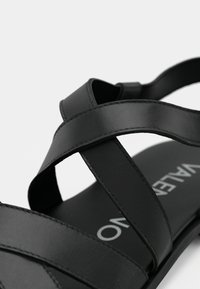 Czarna sandał z szerokimi, krzyżującymi się paskami z gładkiego materiału syntetycznego. Wnętrze pokazuje nazwę marki białym tekstem.