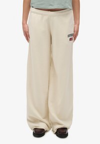 Beige wijde sweatpants met een gladde textuur, elastische tailleband en een klein geborduurd logo op de voorkant links. Aangepaste pasvorm bij de enkels.
