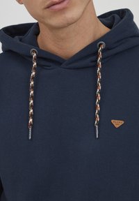 Hoodie in Marineblau aus weichem Stoff, mit einer geflochtenen Kordel mit orange, weiß und schwarzen Akzenten sowie einem kleinen Leder-Logo-Patch.