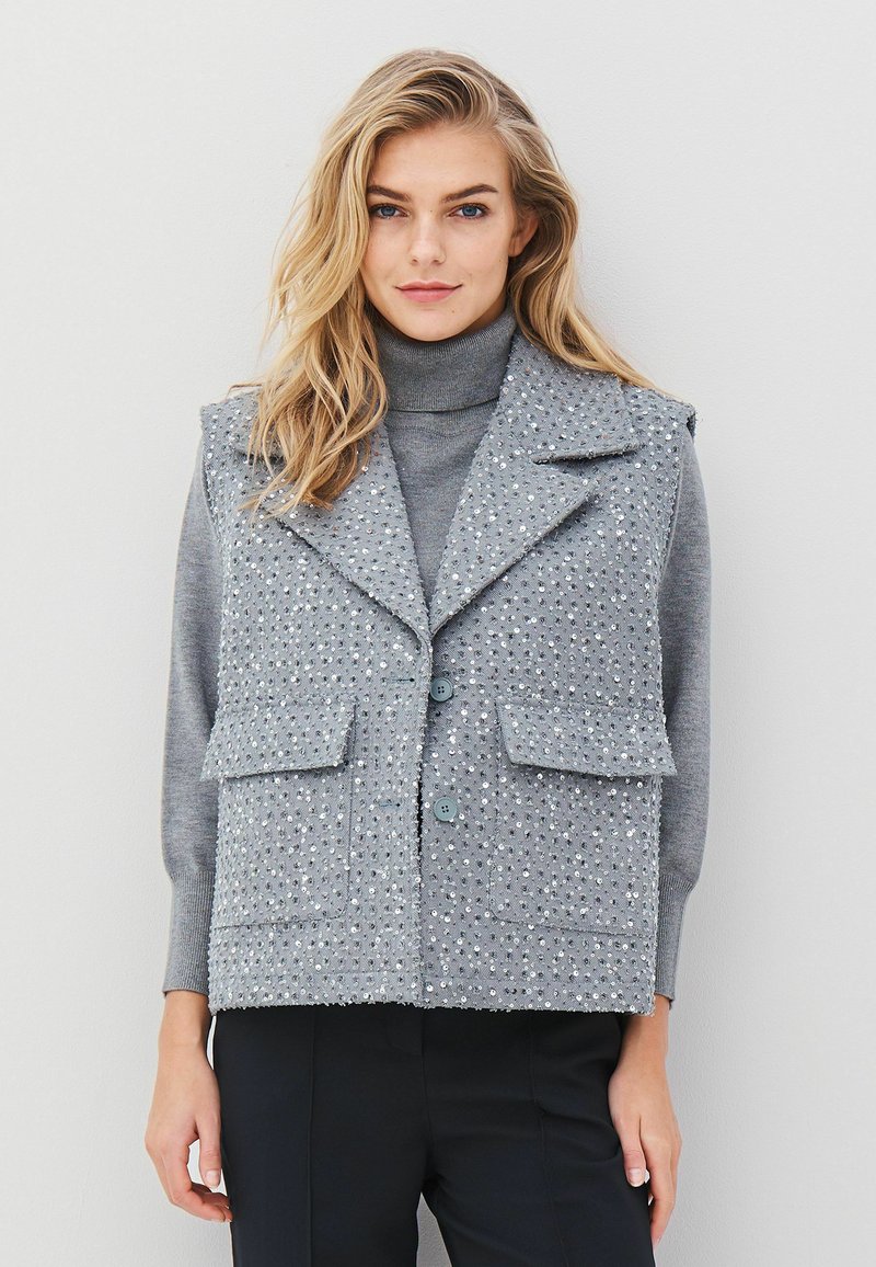 Blonde woman wearing a grey turtleneck sweater under a sequined, buttoned grey vest with large lapels and black pants.