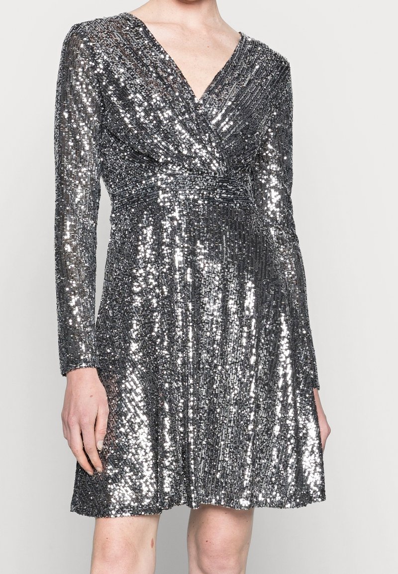 Femme portant une robe pailletée argentée mi-longue, manches longues, col en V et taille froncée.
