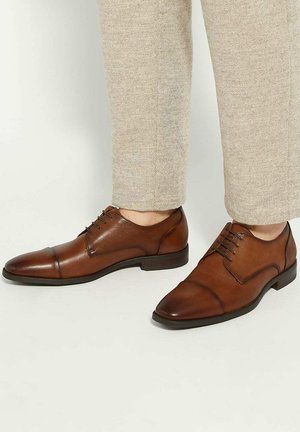 Bruine leren cap-toe oxford schoenen gedragen met beige gestructureerde broek tegen een effen lichte achtergrond.