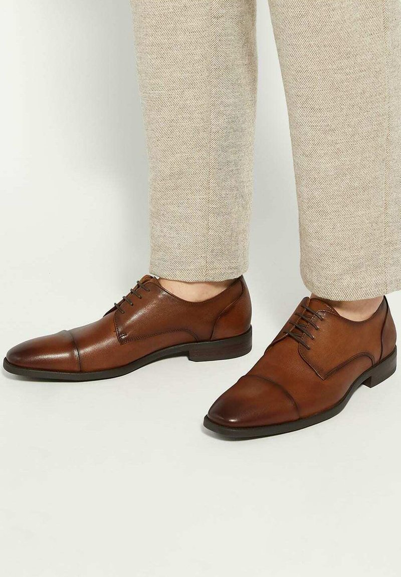 Brown leather cap-toe oxford shoes worn with beige textured trousers against a plain light background.
