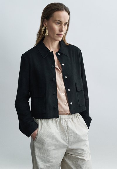 Μαύρο cropped σακάκι από σουέντ με γιακά, μπροστινές τσέπες και ασημένια κουμπιά, φορεμένο πάνω από ένα ανοιχτό μπεζ πουκάμισο και ριγέ παντελόνια.