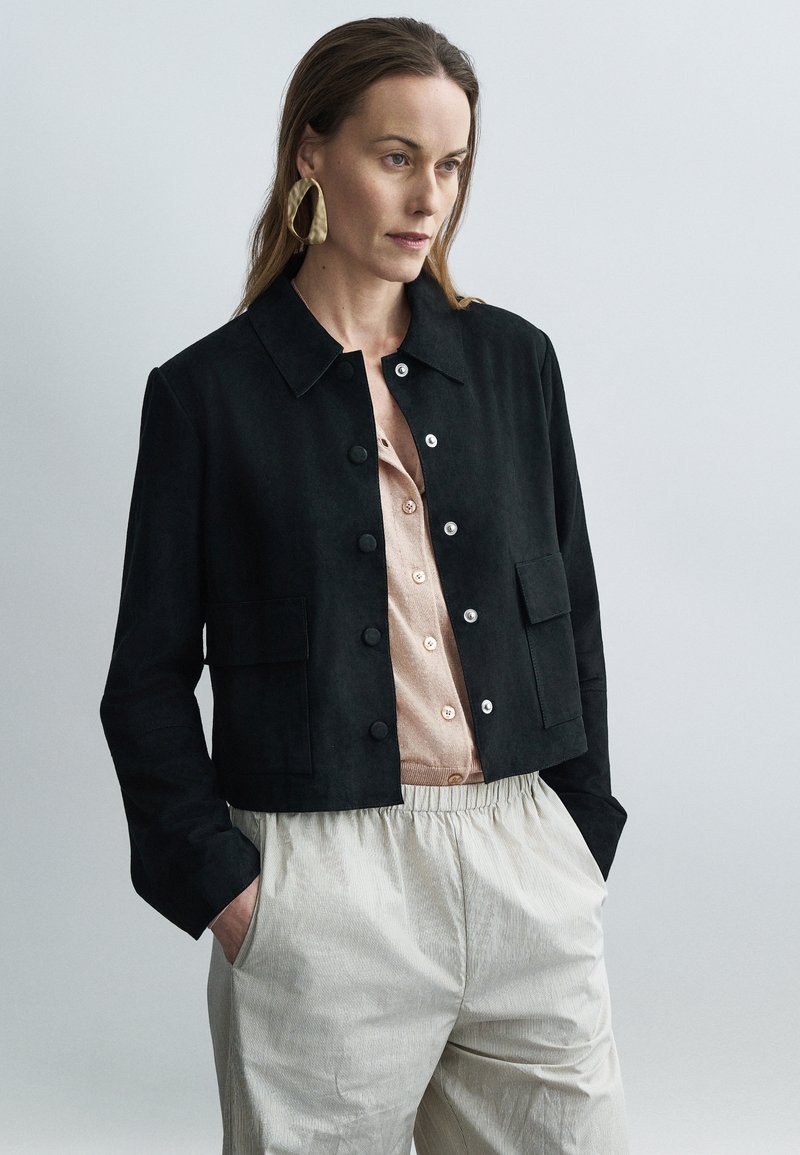 Μαύρο cropped σακάκι από σουέντ με γιακά, μπροστινές τσέπες και ασημένια κουμπιά, φορεμένο πάνω από ένα ανοιχτό μπεζ πουκάμισο και ριγέ παντελόνια.