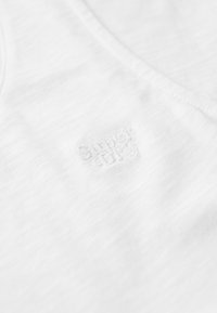 Witte stof met een subtiele textuur. Bevat een verlopen logo in de rechterbenedenhoek, met een v-hals ontwerp en zonder zichtbare patronen.