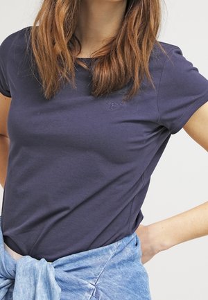 Vrouw met middelbruin haar die een marineblauw T-shirt en een lichtblauw overhemd om haar middel geknoopt draagt, tegen een effen achtergrond.