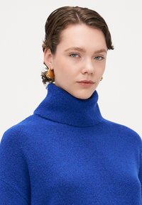 Pulover albastru cu guler înalt dintr-un material tricotat texturat. Cercei geometrici aurii remarcabili.