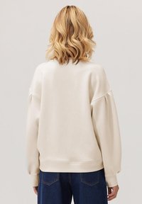 Blondyn z falami do ramion, ubrany w oversize'owy kremowy sweatshirt i ciemnoniebieskie dżinsy, zwrócony plecami na jednolitym tle.