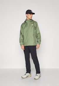 Veste imperméable verte avec fermeture éclair à l'avant, poignets élastiques et accent logo. Portée avec un pantalon noir et des baskets légères multicolores.