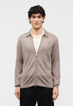 Ung mand med krøllet hår iført beige cardigan med lynlås over hvid skjorte og sorte bukser, stående mod en ensfarvet hvid baggrund.