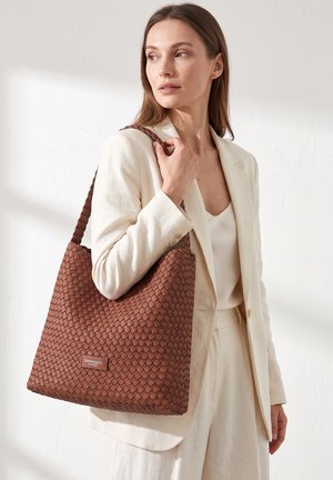 Femme en costume crème portant un grand sac à main en cuir tressé marron avec une bandoulière tressée et une étiquette "DIANA&CO FIRENZE".